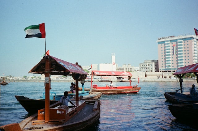Traditional wooden abra boats ferrying passengers across Dubai Creek in the UAE, with the iconic UAE flag waving proudly and modern high-rise buildings in the background – a classic scene of old and new Dubai blending seamlessly.